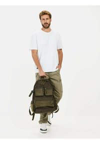 Mørkegrøn stofrygsæk med flere lommer, lynlåse og justerbare stropper, holdt af en person iført en hvid T-shirt og beige bukser.