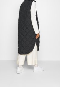 Schwarze gesteppte lange Weste mit hohem Kragen und seitlichen Schlitzen, getragen über einem weißen strukturierten Pullover und cremefarbenen weit geschnittenen Hosen. Weiße Sneakers.