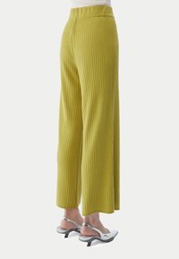 Limegröna ribbade vida byxor kombinerade med en vit topp och silverfärgade slingbackklackar på en stående person, sedd från sidan.