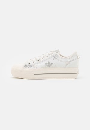 Białe niskie trampki z materiału typu canvas z wiązaniem na przodzie, grubą gumową podeszwą z bocznym paskiem oraz subtelnym wzorem logo Adidas na cholewce.