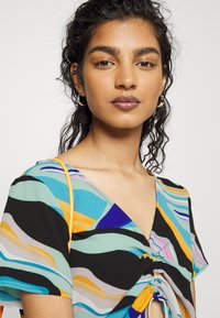 Top de mangas curtas com padrão ondulado multicolorido em azul, preto e laranja; apresenta um detalhe de laço na parte da frente e uma alça laranja.