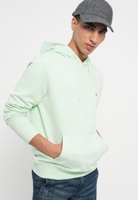 Mintgrønn hettegenser med frontlomme og snor i hetta, laget av mykt stoff. Kombinert med denimjeans og en grå fiskebein-cap.