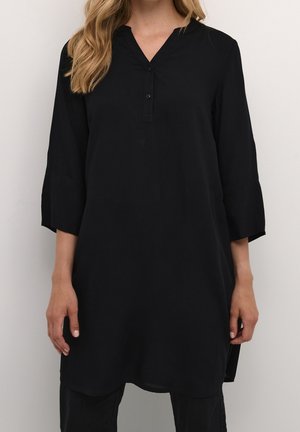 Kobieta w luźnej czarnej tunice z rękawami do łokcia i częściowym zapięciem na guziki, stojąca na tle gładkiego jasnego tła.