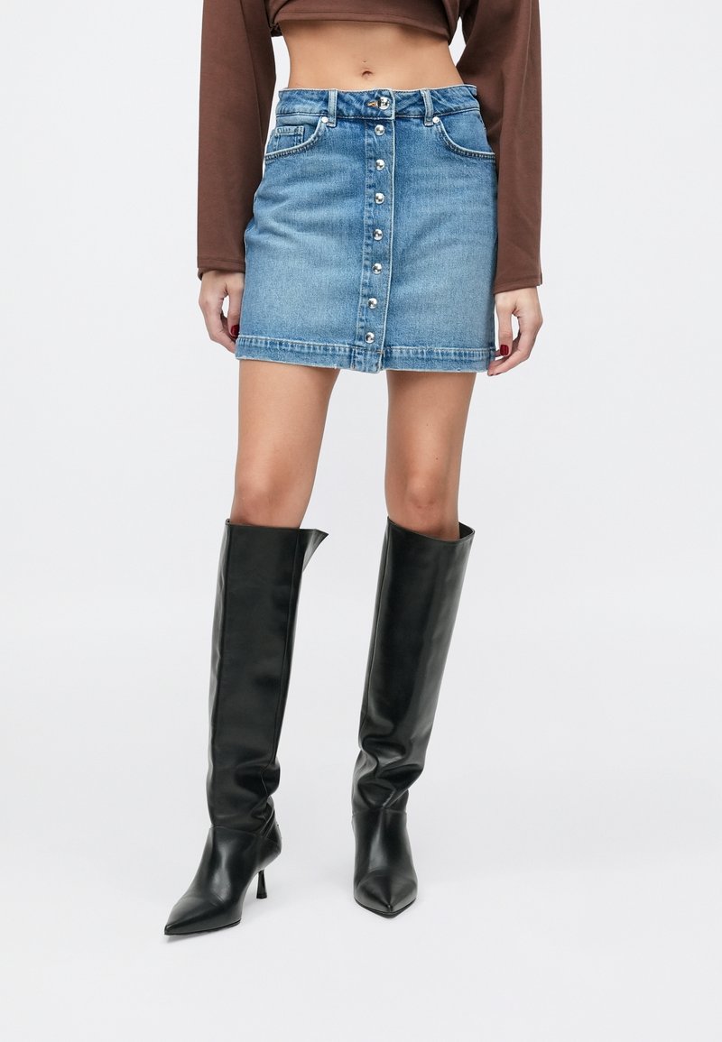 Femme portant une mini-jupe en denim bleue à boutons sur le devant, un haut court marron à manches longues et des bottes noires à talons montant jusqu'au genou.