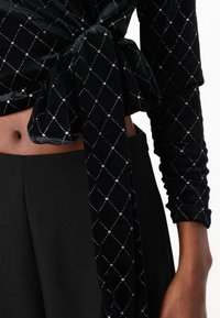 Μαύρο βελούδινο τοπ με δέσιμο μπροστά. Διαθέτει διαμαντένιο σχέδιο διακοσμημένο με ασημένιες τρύπες. Μακριά μανίκια, λεία υφή και εφαρμοστή σχεδίαση.