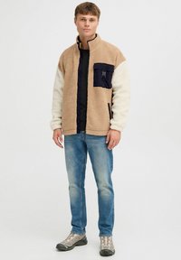 Chaqueta de forro polar con secciones en color marrón claro, crema y marino; cuello alto y cierre con cremallera; cuenta con un bolsillo en el pecho con clip; llevada sobre una camisa negra.