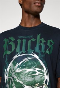 Marineblå T-shirt med teksten "Milwaukee Bucks" i grøn. Centergrafik viser en stiliseret basketball med sølvaccentuer og et tekstureret mønster.