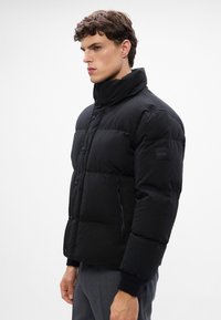 Jachetă puffer neagră cu guler înalt, închidere cu nasturi și buzunare laterale cu fermoar. Prezintă secțiuni matlasate și o P patch logo pe mânecă.
