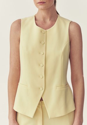Ärmellose, blassgelbe Weste mit Knöpfen und zwei Vorder Taschen, getragen über passenden Hosen an einer Person vor neutralem Hintergrund.