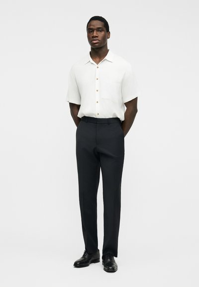 Homme debout, les mains dans le dos, portant une chemise blanche à manches courtes boutonnée, un pantalon de costume noir et des chaussures de ville noires.