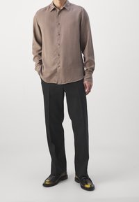 Camisa marrón claro de manga larga con cuello abotonado, cierre de botones y mangas con puños, combinada con pantalones negros y zapatos brillantes.