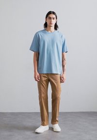 Camiseta de algodón de manga corta en azul claro combinada con pantalones marrones texturizados y zapatillas blancas, modelada por una persona con pelo largo.