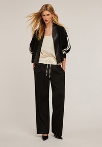 Svart kort bomberjacka med vita ränder, krämfärgad satinblus och lösa svarta byxor. Har en dragsko i midjan och eleganta klackar.