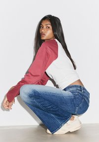 Jeune femme aux longs cheveux foncés, portant un t-shirt à manches longues rouge et blanc, un jean bleu et des baskets beige, accroupie contre un mur blanc.