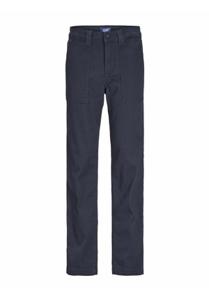 Dziecko ubrane w granatową koszulkę polo z logo, granatowe spodnie z kieszeniami oraz białe trampki, stojące na tle jednolitego jasnego tła.