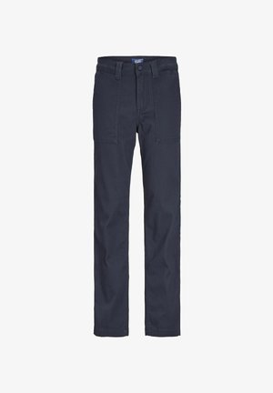 Dziecko ubrane w granatową koszulkę polo z logo, granatowe spodnie z kieszeniami oraz białe trampki, stojące na tle jednolitego jasnego tła.