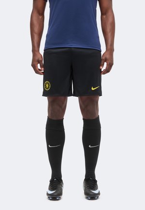 Mandlig atlet iført sorte Chelsea fodboldshorts, sorte Nike sokker, sorte støvler og en mørkeblå skjorte, stående oprejst mod en hvid baggrund.