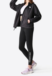 Giacca nera leggera con zip, tasche laterali e polsini elasticizzati; abbinata a leggings neri con branding bianco e sneakers lilla.