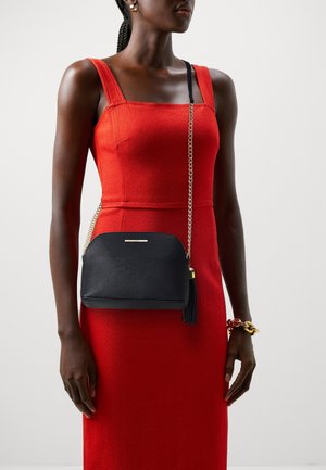 Woman wearing a sleeveless red dress and gold bracelet, carrying a black crossbody bag with a gold chain strap and tassel.