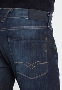 Mörkblå denimjeans har en standard femfickdesign med en logotyp-patch på midjan och kontrasterande sömmar på bakfickorna.