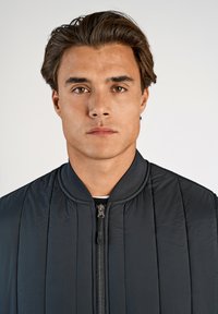 Svart quiltad bomberjacka med ribbad krage och dragkedjestängning. Slät textur, figurnära design med vertikalt sydda linjer.