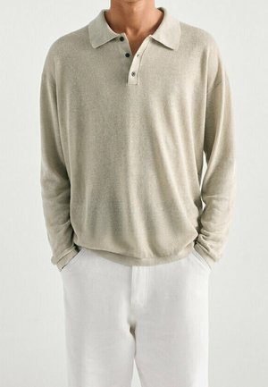 Homme portant un polo beige à manches longues avec trois boutons et un pantalon blanc, mains dans les poches, sur un fond clair uni.