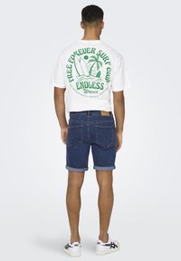 T-shirt bianca con stampa grafica verde sulla schiena, abbinata a pantaloni shorts in denim blu e sneakers bianche. Visibili i risvolti sui pantaloni.