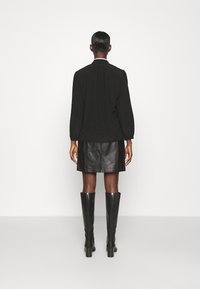 Schwarzes Langarmoberteil mit hohem Kragen, kombiniert mit einem schwarzen Leder-Minirock und knielangen, hochhackigen Stiefeln, von hinten gezeigt.