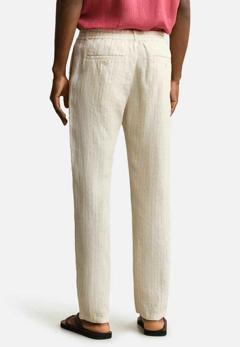 Vista trasera de una persona que lleva pantalones beige con rayas finas, camisa roja y chanclas negras, de pie frente a un fondo claro y sencillo.