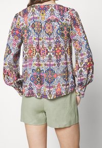 Langarmblouse in een patroonstof met levendige kleuren zoals roze, blauw en groen, met plooidetail aan de achterkant. Gecombineerd met groene shorts.