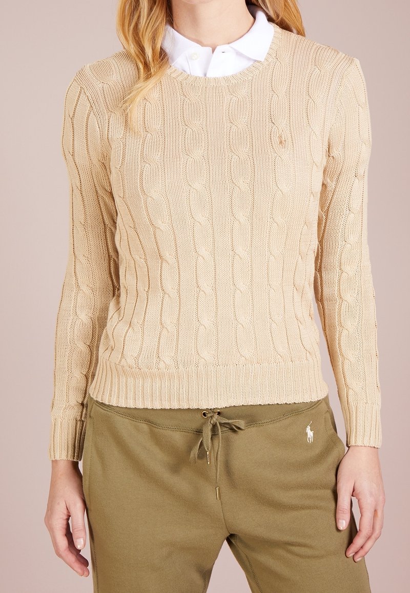 Femme portant un pull en maille torsadée beige sur une chemise blanche à col et un pantalon à cordon de serrage vert olive avec logos brodés.