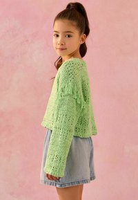Grüne gehäkelte Bluse mit Fransen, langen Ärmeln und offenem Strickmuster, kombiniert mit Jeansshorts. Der Hintergrund ist rosa.
