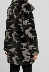 Abrigo de camuflaje difuso con una mezcla de tonos verde oscuro y gris, con cuello tipo solapa y un corte relajado. Textura suave y mangas largas.