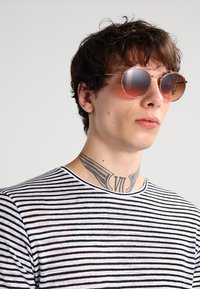 Okuliare s okrúhlym rámom a hnedými gradientovými sklami, tenkým kovovým rámom a štíhlym dizajnom. Model má na sebe prúžkované čierne a biele tričko.
