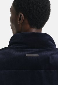 Navy corduroy jas met een hoge kraag, ribbeltextuur en een klein metalen merkteken op de achterkant.
