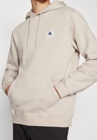 Beige hoodie i mjukt material med känguruficka, dragsko huva och en cirkulär logotyp med en blå stjärna.