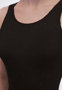 Κοντινό πλάνο ενός ατόμου που φοράει μαύρο, ριγωτό, αμάνικο τοπ με στρογγυλεμένη λαιμόκοψη σε ουδέτερο φόντο.