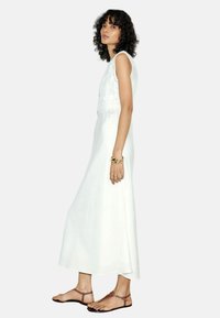 Robe maxi sans manches blanche avec une finition texturée, une silhouette fluide et une fente latérale. Modèle portant des sandales plates marron et des bijoux en or.