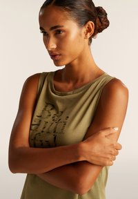 Top senza maniche verde oliva realizzato in un tessuto morbido, con un design testuale nero decorato con borchie, scollatura rotonda e vestibilità ampia.