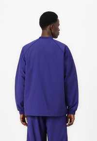 Veste en nylon violet avec une coupe droite, des manches raglan et des poignets élastiques. Présente un design simple, sans logos ni motifs visibles.