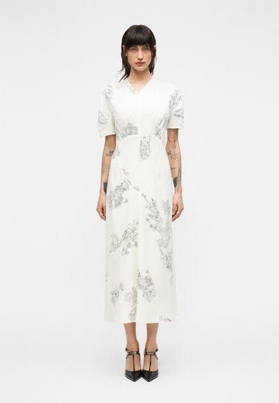 Kvinde med mørkt hår iført en hvid midi-kjole med korte ærmer og sorte blomsterprint, kombineret med sorte T-rem hæle.