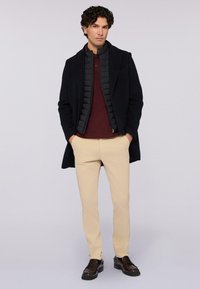 Muž oblečený v černém kabátu přes černou prošívanou bundu, vínové polo tričko, béžové kalhoty a tmavě hnědé společenské boty, stojící s rukama v kapsách.