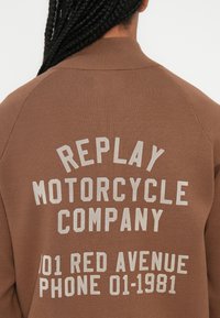 Brun genser med teksten "REPLAY MOTORCYCLE COMPANY 101 RED AVENUE TELEFON 01-1981" i lysegrå, med riller.