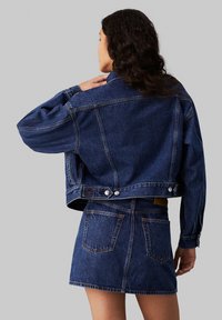 Chaqueta de mezclilla con un diseño corto, color azul, y detalles de botones en la cintura, combinada con una falda de mezclilla a juego que cuenta con bolsillos en la parte trasera.