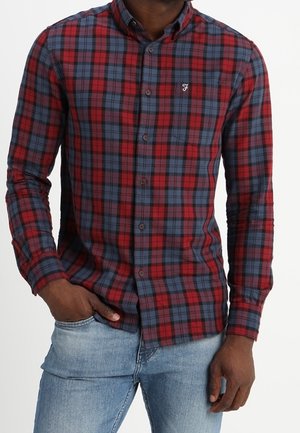 Hombre con una camisa de cuadros roja y azul con botones y mangas largas y vaqueros azul claro, de pie con una mano en el bolsillo.