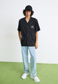Svart t-skjorte med kort erme og logo, med en enkelt rad med knapper, kombinert med lyseblå jeans og hvite sneakers, plassert på et grønt gulv.