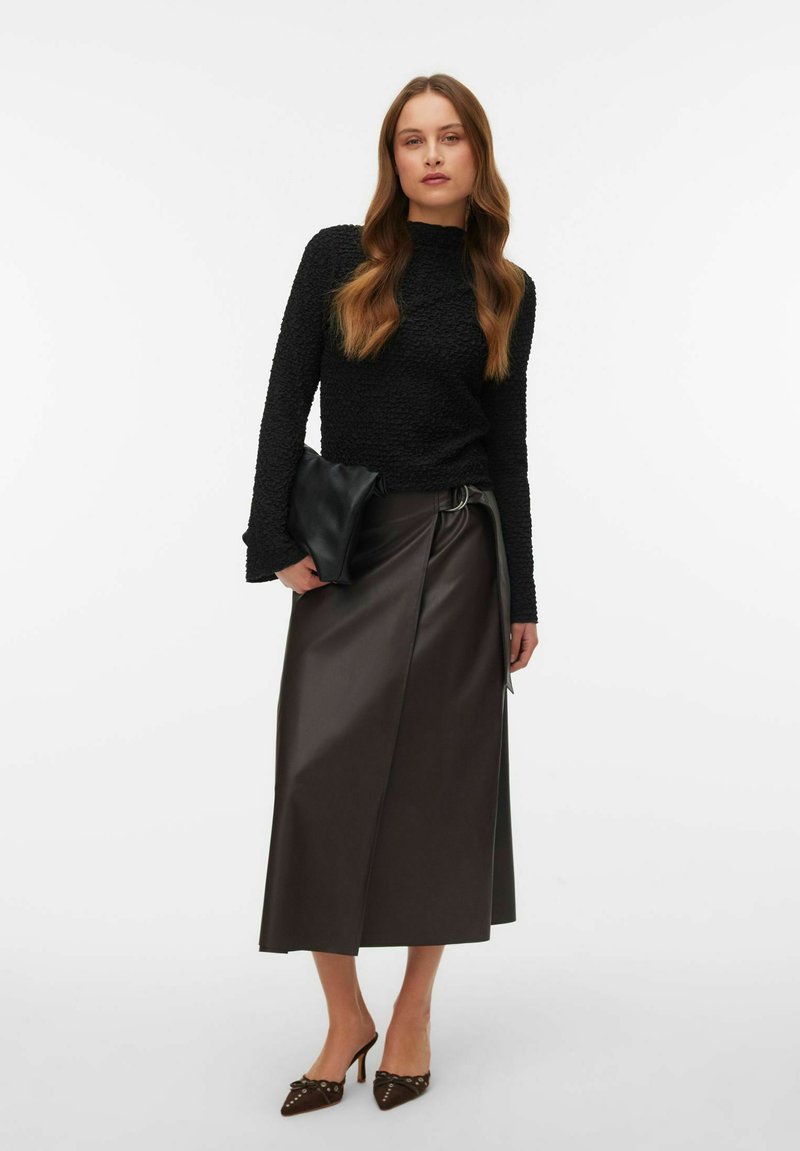 Vrouw draagt een zwart, gestructureerd, langärmelig shirt, een midi-rok van bruin leer met riem, bruine pumps met studs, en houdt een zwarte clutch vast.