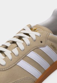 Μπεζ αθλητικό παπούτσι Adidas Samba με λευκά κορδόνια, τρεις λευκές ρίγες στο πλάι και καφετί σόλα, εμφανίζοντας κοντινή λήψη των λεπτομερειών των κορδονιών και του λογότυπου.