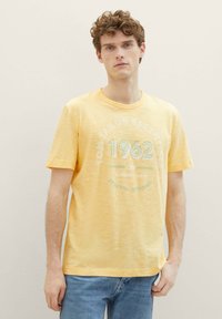 Camiseta amarilla de manga corta con un acabado texturizado, escote redondo y texto impreso que dice "TOM TAILOR ESSENTIALS 1962."