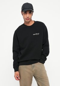 Melna sporta krekla ar ribotām piedurknēm un apakšmalu, uz krūtīm ir neliels balts logotips. Saskaņots ar gaiši brūnām biksēm un melnu cepuri.